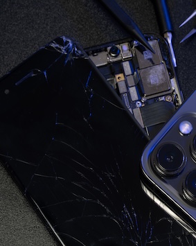 Shattered smartphone with exposed internal components on a dark surface, illustrating repair.