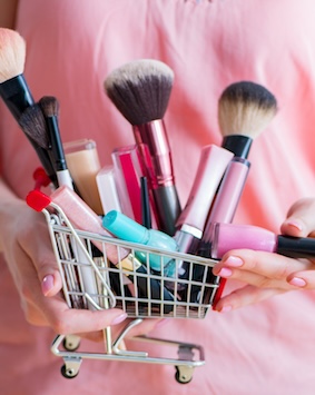 A miniature shopping cart filled with various makeup products held in a person's hand.