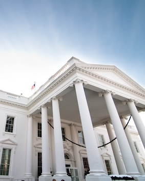 Upward view of the White House's neoclassical facade with its iconic columns and portico, under a clear sky.