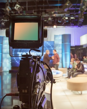A television studio camera in focus, with a blurred background showing hosts and the set.