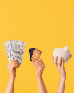 Image showing three hands raised against a yellow background, one holding cash, another holding credit cards, and the third holding a piggy bank.